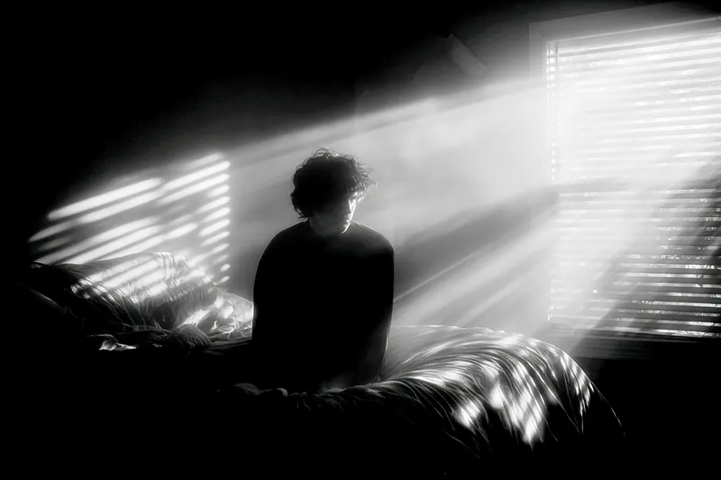 A figure sitting at the edge of their bed at 2 AM, moonlight streaming through blinds, creating patterns of light and shadow